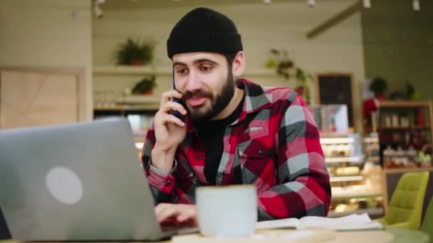 Moderner Coffeeshop attraktiver Kerl unterhalten sich auf seinem Smartphone, während er online am Laptop arbeitet, er hat eine Tasse Kaffee auf dem Tisch — Stockvideo