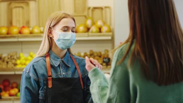 Koronavirüs salgınında koruyucu maskeli satıcı kadın organik sebze dükkanına girmeden önce müşterinin sıcaklığını kontrol eder. — Stok video