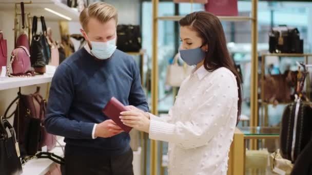 Charismatic mature saleswoman try to help the customer man to choose a right present for some special person from the accessories shop in the pandemic of COVID 2019 they wearing protective masks — Stock Video