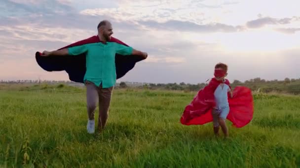 Przystojny, charyzmatyczny chłopczyk i jego ojciec biegający po zielonym polu w garniturach i maskach superbohaterów uśmiechają się i czują się szczęśliwi. — Wideo stockowe