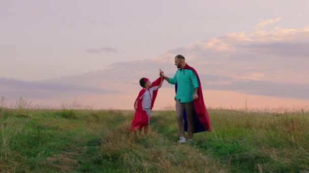 Saat matahari terbenam ayah dan anaknya membuat tim yang sulit bersama-sama mereka mengenakan setelan superhero berjalan di depan kamera melalui lapangan hijau — Stok Video