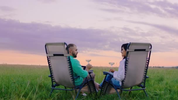 Niesamowita para cieszy się chwilą na łonie natury patrząc w niebo o zachodzie słońca siedząc na krzesłach na środku zielonego pola. 4k — Wideo stockowe
