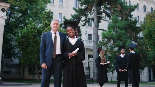 Journée de graduation charismatique dame noire diplômée posant avec le professeur d'université devant la caméra tout en tenant le diplôme — Video