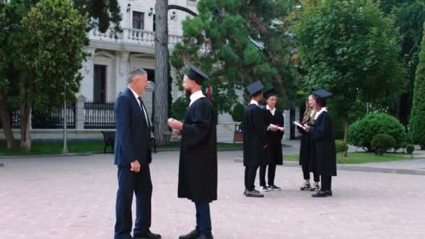 Ευτυχισμένος μαθητής συζητά με τον πατέρα του μετά την αποφοίτηση πολύ ενθουσιασμένος που κατέχει το πτυχίο του και φοράει καπέλο και κοστούμι αποφοίτησης — Αρχείο Βίντεο