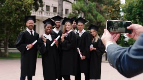 Día de graduación en el grupo del parque universitario de graduados multirraciales estudiantes posando emocionados con diplomas delante del teléfono inteligente el director de la universidad tomando fotos — Vídeos de Stock