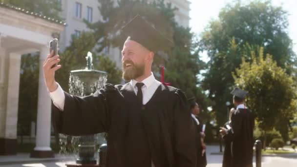 Hombre guapo con una barbilla graduado en el jardín de la universidad tomar su teléfono inteligente para hacer una llamada con la cámara que muestra su diploma y luego seguir hablando en el teléfono inteligente — Vídeos de Stock