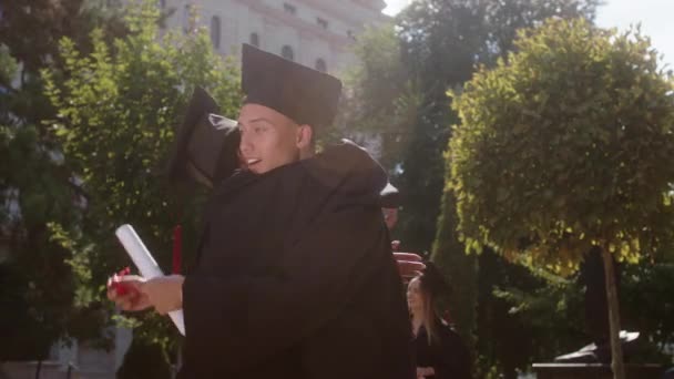 No jardim da faculdade após o dia de formatura dois estudantes multirraciais senhora e cara abraçando uns aos outros depois de tomar os diplomas — Vídeo de Stock