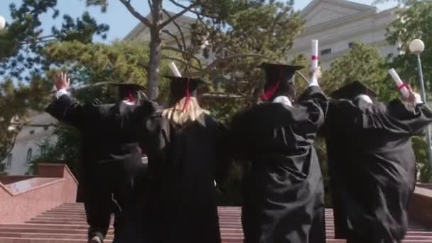Heyecanlı, çok kültürlü bir grup öğrenci üniversite binasının merdivenlerinden koşarak çıkıyorlar. Mezuniyet şapkaları takıyorlar ve diplomaları ellerinde tutuyorlar. — Stok video