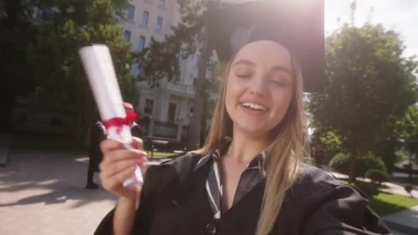 在一个阳光普照的完美日子里，漂亮的毕业生兴奋极了，为她的父母制作了一个自拍视频，她在大学校园里拿着毕业证书说话 — 图库视频影像