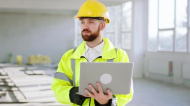 İnşaat alanında kameraya doğru yürürken aynı zamanda dizüstü bilgisayarda çalışırken aynı zamanda iyi görünümlü bir mühendis elinde dizüstü bilgisayarla inşaat planını analiz ediyor.