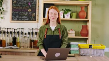 Kahve dükkanında çalışan genç bayan barista dizüstü bilgisayarın başında duruyor bir şeyler yazıyor ve doğrudan kameraya bakıyor ve kocaman gülümsüyor.