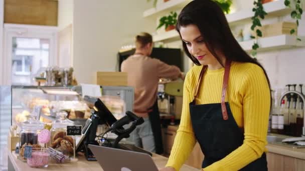 Beautiful woman working barista in the coffee shop she typing a online order on the laptop while other guy barista start to do the new order — Stock Video