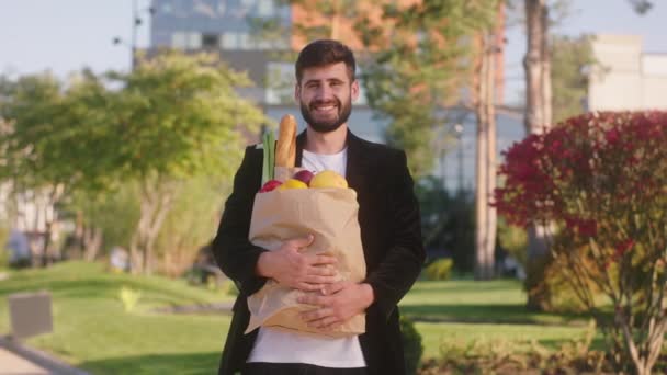 An einem sonnigen Tag in der Mitte der modernen Straße posiert der charismatische Mann mit einem perfekten Lächeln vor der Kamera und hält eine Öko-Einkaufstasche voller Lebensmittel und Gemüse in der Hand. Arri Alexa