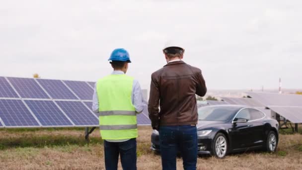 現代の太陽電池パネルの概念大規模太陽電池を歩く2人の生態技術者は、デジタルタブレットを使用して一緒に議論し、保護ヘルメットを身に着けている — ストック動画