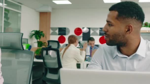 Um homem negro misto atraente e uma mulher branca estão no trabalho conversando, no fundo dois homens bonitos estão olhando para um tablet e sorrindo alegremente — Vídeo de Stock