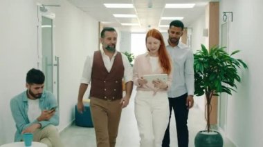 Üç iş arkadaşı ofisteki bir masaya birlikte yürüyorlar, yürürken insanları selamlıyorlar, masada otururken iş hakkında konuşuyorlar.