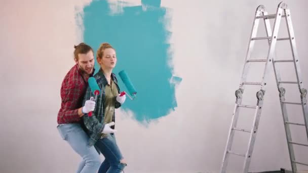 Pareja feliz y divertida mientras pintan las paredes usando el rodillo de cepillo de pared para cantar juntos divertirse y disfrutar del momento — Vídeo de stock