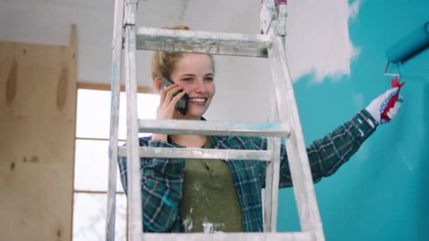 Charismatische vrouw tijdens het schilderen van de muren thuis in een blauwe kleur spreekt op haar smartphone met iemand en met een grote glimlach — Stockvideo