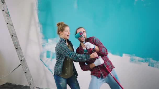 Em casa na sala de estar casal feliz e animado ao pintar as paredes dançando e cantando usando as escovas para que eles têm um tempo perfeito juntos — Vídeo de Stock