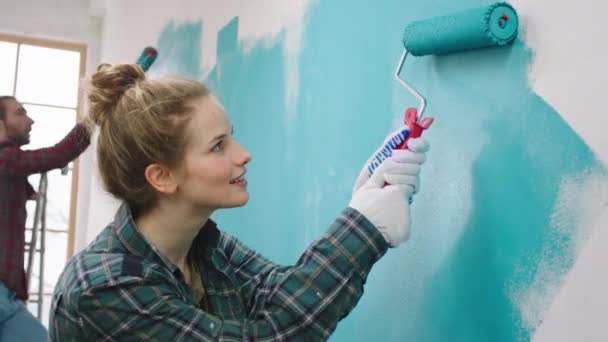 Charismatic cu o femeie zâmbet frumos și soțul ei acasă care lucrează împreună pictura pereți în culoarea albastră folosind pensula de perete — Videoclip de stoc
