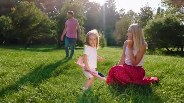 In einem großen grünen Park genießen glückliche Familie mit einem netten kleinen Mädchen die Zeit zusammen genießen die Sonne und spielen mit einem rosa Flugzeug — Stockvideo