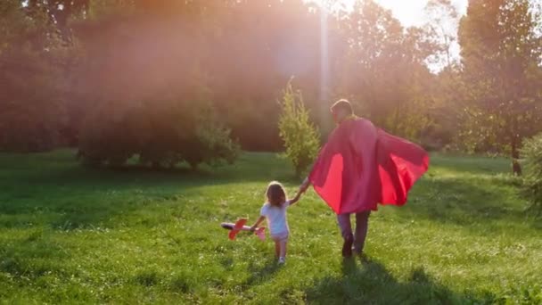 Perfect zonnige dag voor een vader en zijn kleine dochter die samen spelen in het midden van het park ze lopen door het gras de vader draagt een superheld pak — Stockvideo