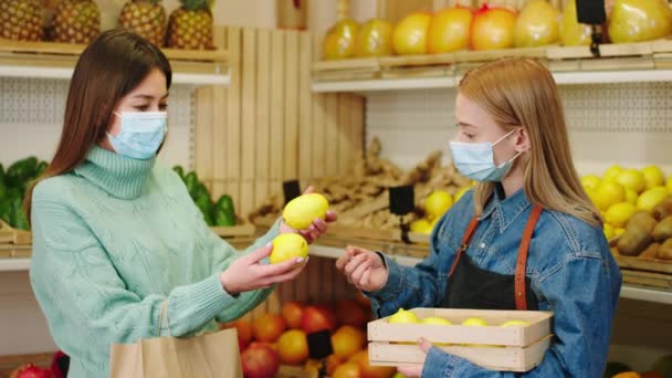 В пандемии коронавируса в органическом овощном магазине продавщица держит сундук с спелыми лимонными фруктами и помогает женщине-клиенту взять несколько хороших фруктов они носят маску — стоковое видео