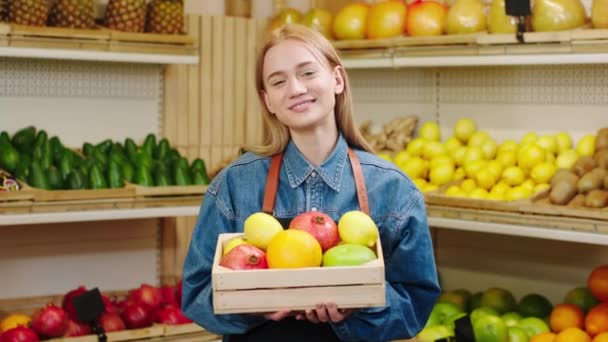 W organicznym sklepie warzywnym niesamowita młoda kobieta rolnik trzyma klatkę piersiową pełną warzyw i pozowanie przed kamerą z dużym uśmiechem — Wideo stockowe