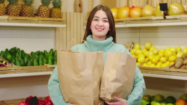 Voor de camera ziet er goed uitziende dame met een grote glimlach in de biologische groentewinkel met eco zakken vol verse rijpe groenten en fruit — Stockvideo