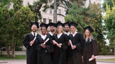 Modern üniversite parkındaki çok ırklı mezunlar mezuniyetten sonra ellerinde diplomalarla kameraların önünde poz veriyorlar.