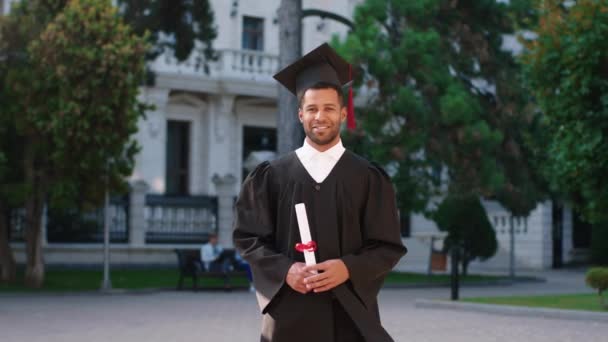 Красивий чорний хлопець випускник позує перед камерою зі своїм дипломом посміхаючись великим і відчуваючи збудження в день випуску — стокове відео