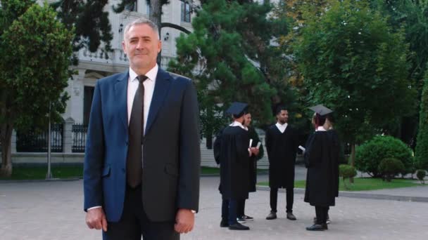 Üniversite parkında kamera önünde mezuniyet konsepti. Takım elbiseli karizmatik bir adam kameranın önünde poz veriyor. Arka plandaki öğrencilere bakıyor. — Stok video