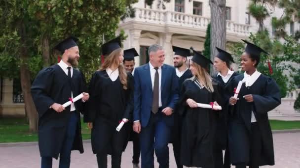 Wieloetniczni absolwenci w parku studenckim spacerujący w grupie z dyrektorem koledżu po ukończeniu szkoły bardzo ekscytująco trzymający dyplomy i dyskutujący razem — Wideo stockowe