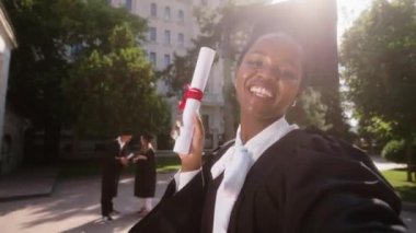 Mezuniyet gününde büyük bir üniversite bahçesi. Siyahi güzel bir mezun heyacanlı. Kamerayla bir selfie videosu çek.