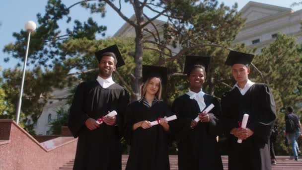 Grupo de graduados multirraciais estudantes na frente da câmera posando animado a formatura que detém diplomas e bonés de graduação e ternos — Vídeo de Stock