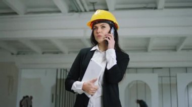 Kameranın önünde, karizmatik denetçi kadın dijital tabletini tutarken ve güvenlik kaskı takarken akıllı telefonuyla sohbet ediyor.