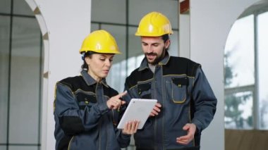 İnşaat alanında bir kadın mühendis ve inşaat işçisi dijital tablet kullanarak inşaat planını analiz ediyor ve birlikte tartışıyorlar.