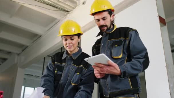 Feliz y entusiasta dos trabajadores de la construcción mujer y hombre utilizando tableta digital y algunos papeles para analizar el plan de construcción que llevan equipo de protección y cascos. — Vídeos de Stock