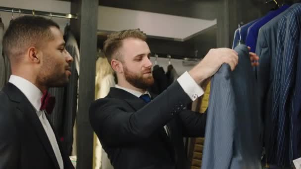 Brudgommen er at vælge det rigtige jakkesæt til brylluppet i dragtbutikken han har en diskussion med konsulenten karismatisk mand – Stock-video
