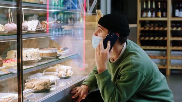 Delante de la cámara en la cafetería en la pandemia de coronavirus hombre cliente con máscara protectora elegir un postre de la nevera escaparate mientras habla en el teléfono inteligente — Vídeos de Stock