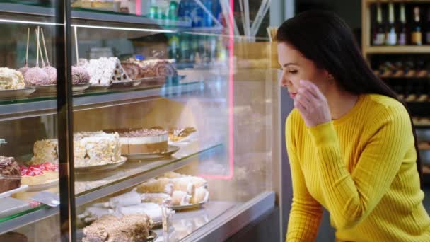 Tomando detalles en el café de la panadería de una hermosa mujer cliente mirando la nevera escaparate lleno de desiertos frescos para elegir el favorito — Vídeos de Stock