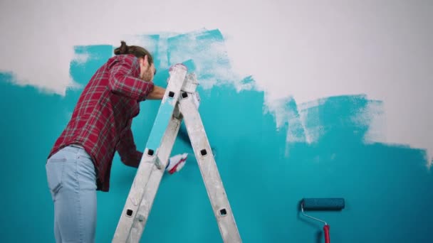 Delante de la cámara en la escala, la pintura del hombre concentró las paredes en el color azul usando el rodillo de cepillo de pared. Se cansa al final. — Vídeos de Stock