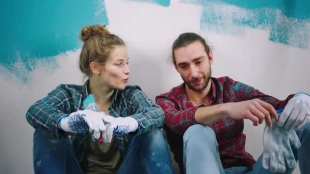 Kaukasisches Paar in der Nähe der Kamera diskutiert und lächelt breit, während es eine Pause einlegt, bevor es zu Hause mit dem Bemalen der Wände in blauer Farbe beginnt — Stockvideo