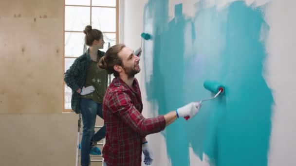 Gelukkig vrouw thuis samen met de man schilderen de muren in de woonkamer ze hebben een grote glimlach met behulp van muur borstel roller — Stockvideo