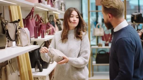 В магазине аксессуаров красивая клиентка смотрит на сумки с полок, в то время как один харизматичный продавец хочет помочь клиенту — стоковое видео