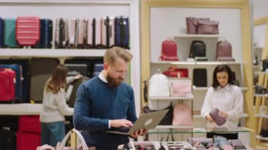 Diğer müşteriler arka plandayken aksesuar dükkanındaki çantaların fiyatını kontrol etmek için dizüstü bilgisayar kullanan yakışıklı bir adam denetçisi.