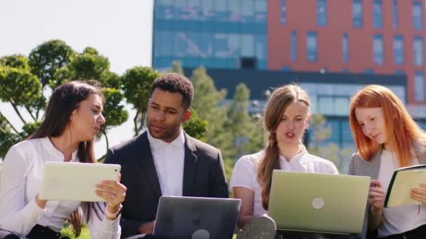 Młodzi studenci odrabiający pracę domową w grupie używając laptopów i tabletów do wspólnej nauki siadają na trawie w nowoczesnym parku studenckim — Wideo stockowe