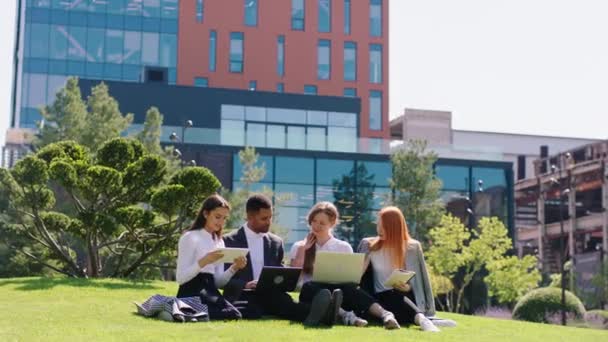 Grupo atraente de estudantes senhoras e um cara preto levar os laptops e tablets para estudar juntos no moderno parque universitário na grama eles ajudam uns aos outros — Vídeo de Stock