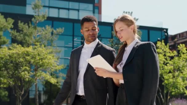 Modern business building architecture happy and excited woman and black guy office managers at the break time walking down the street and using tablet to analysing something — Stock Video