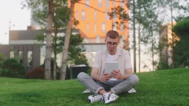 Modern kolej parkındaki gözlüklü genç adam üniversite projesi yapmak için elektronik tablet kullanıyor.